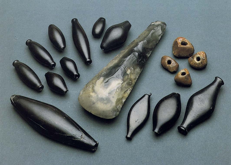 A photograph showing 12 elongated jet beads of various sizes, 4 irregular shaped amber beads, and a polished green marbled flint axe head 